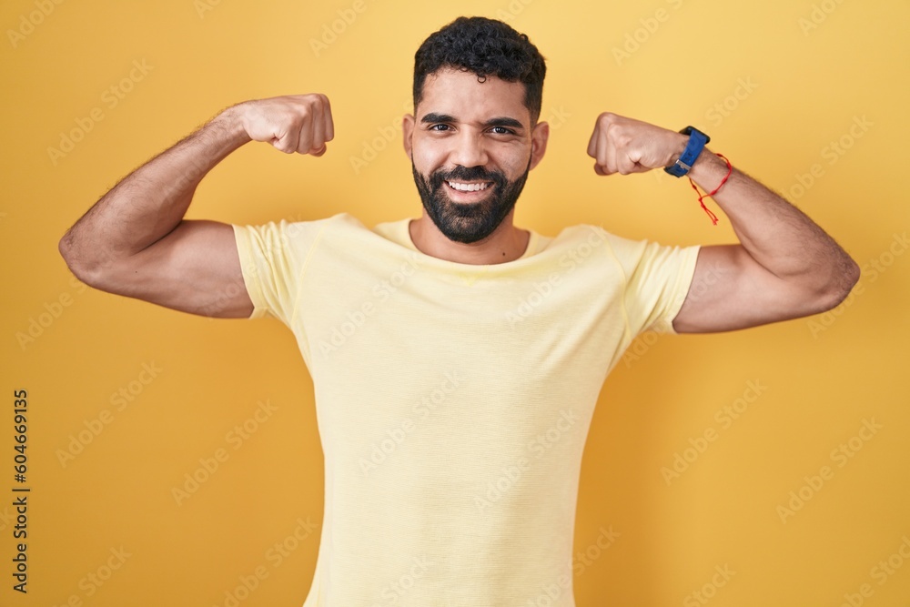 Hispanic man with beard standing over yellow background showing arms muscles smiling proud. fitness concept. Hispanic man with beard standing over yellow background showing arms muscles smiling proud. fitness concept.