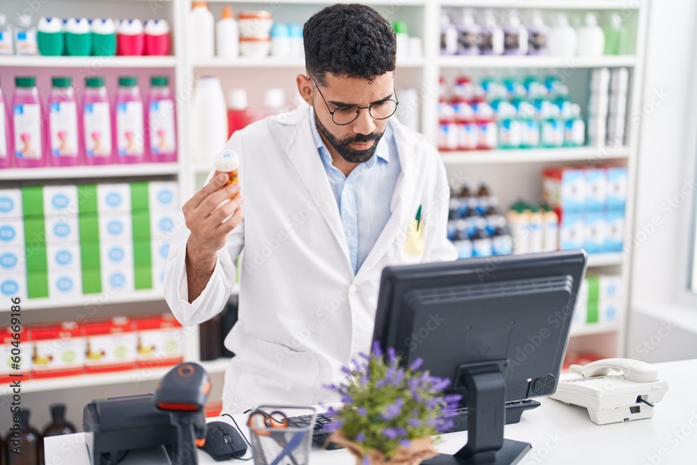 Young arab man pharmacist using computer holding pills bottle at pharmacy