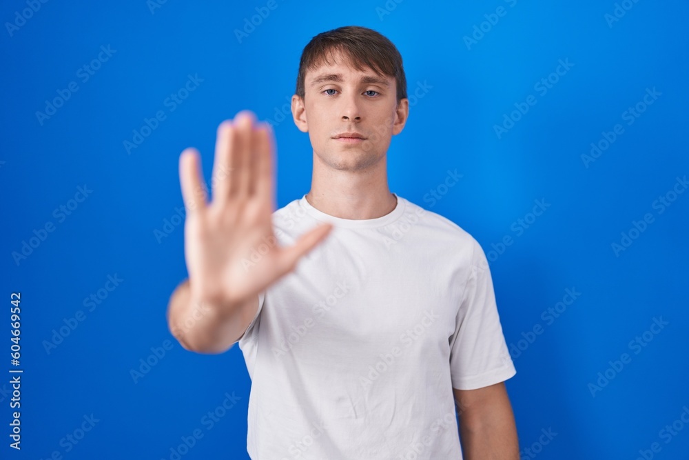 Caucasian blond man standing over blue background doing stop sing with palm of the hand. warning expression with negative and serious gesture on the face. Caucasian blond man standing over blue background doing stop sing with palm of the hand. warning expression with negative and serious gesture on the face.