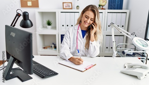 Young woman wearing doctor uniform writing on document talking on smartphone at clinic