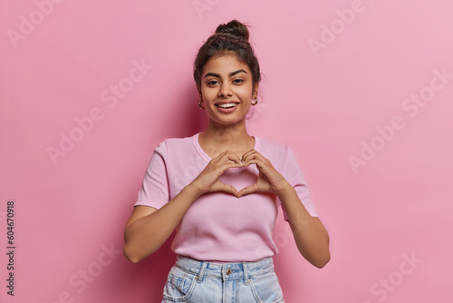 Portrait of happy Indian woman with hair bun shows finger heart gesture sends love to someone smiles pleasantly wears casual clothes isolated on pink background expresses sympathy feels passionate © Wayhome Studio