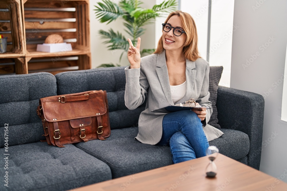 Hispanic woman working at consultation office smiling looking to the camera showing fingers doing victory sign. number two.