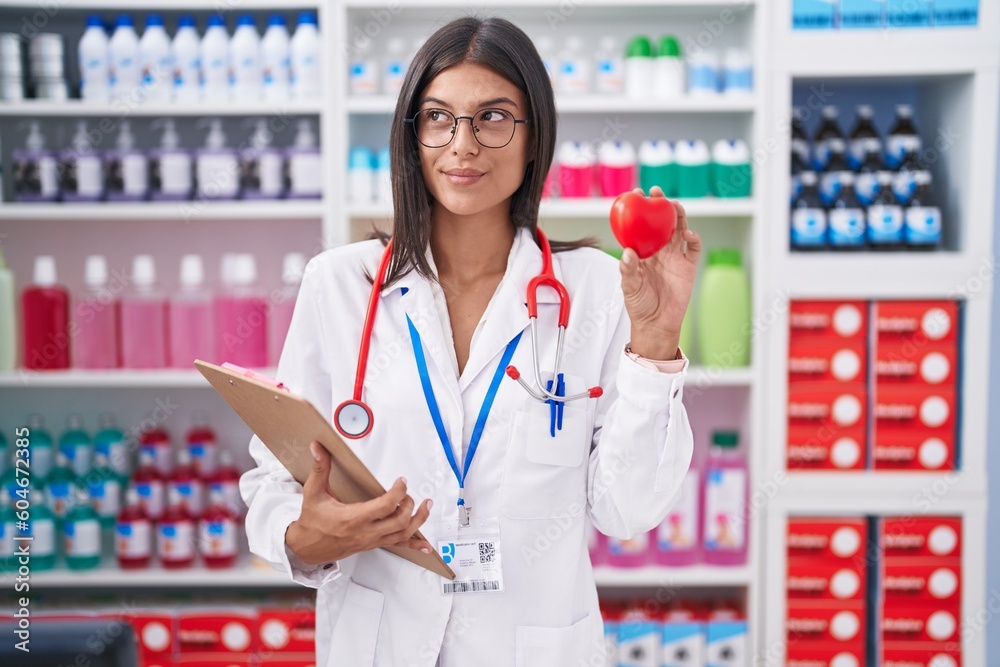 Brunette young woman working at pharmacy drugstore holding red heart smiling looking to the side and staring away thinking.