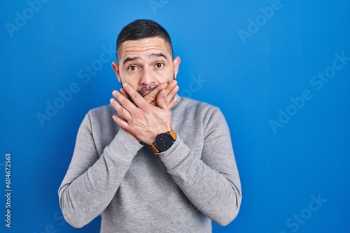 Hispanic man standing over blue background shocked covering mouth with hands for mistake. secret concept.