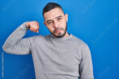 Hispanic man standing over blue background strong person showing arm muscle, confident and proud of power