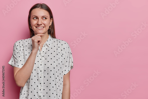Good looking cheerful female model with dark straight hair bites lips keeps hand on chin wears polka dot blouse has dreamy expression isolated over pink background copy space for your advertisement