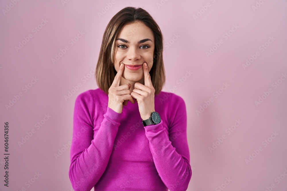 Hispanic woman standing over pink background smiling with open mouth, fingers pointing and forcing cheerful smile Hispanic woman standing over pink background smiling with open mouth, fingers pointing and forcing cheerful smile