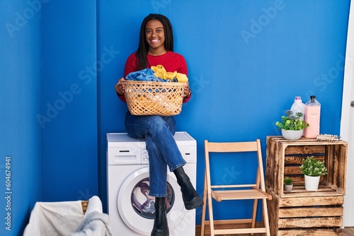 Young african american with braids holding laundry basket sitting on washing machine smiling with a happy and cool smile on face. showing teeth.