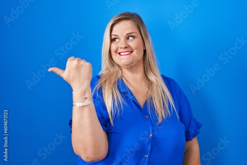 Caucasian plus size woman standing over blue background smiling with happy face looking and pointing to the side with thumb up. © Krakenimages.com