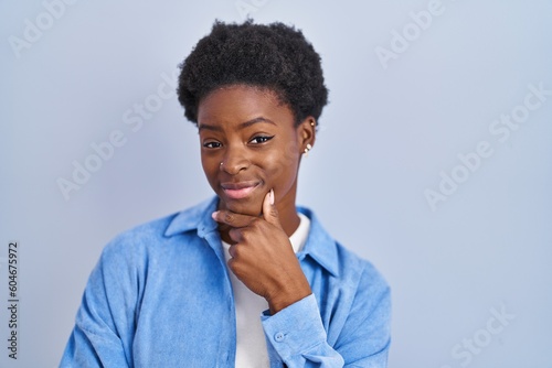 African american woman standing over blue background looking confident at the camera smiling with crossed arms and hand raised on chin. thinking positive.