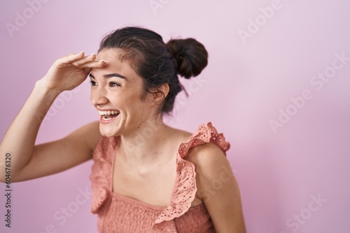 Young teenager girl standing over pink background very happy and smiling looking far away with hand over head. searching concept.