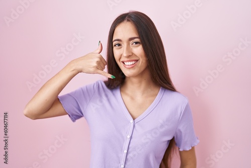 Young hispanic woman with long hair standing over pink background smiling doing phone gesture with hand and fingers like talking on the telephone. communicating concepts.