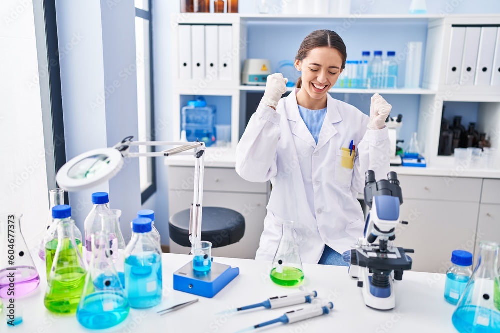 Fototapeta premium Young brunette woman working at scientist laboratory very happy and excited doing winner gesture with arms raised, smiling and screaming for success. celebration concept.