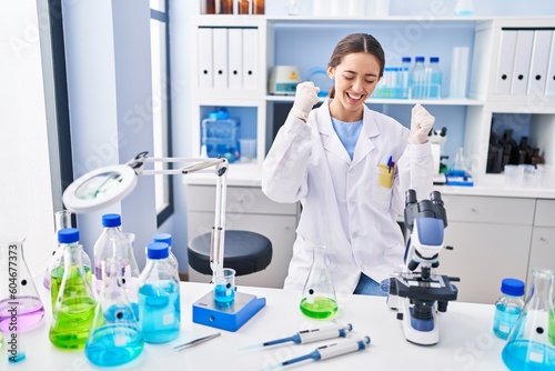 Young brunette woman working at scientist laboratory very happy and excited doing winner gesture with arms raised  smiling and screaming for success. celebration concept.