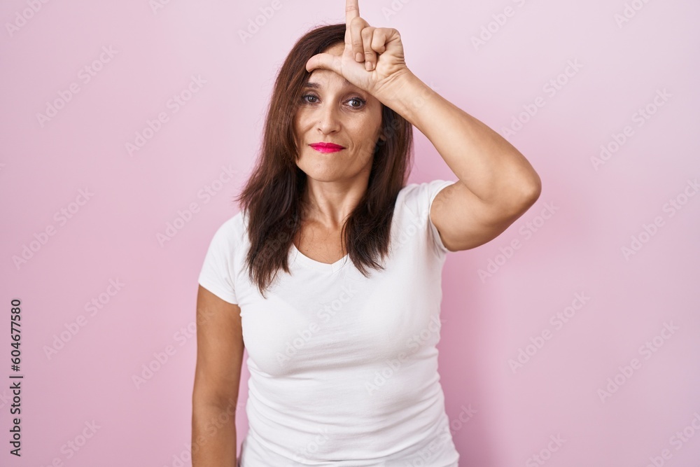 Middle age brunette woman standing over pink background making fun of people with fingers on forehead doing loser gesture mocking and insulting. Middle age brunette woman standing over pink background making fun of people with fingers on forehead doing loser gesture mocking and insulting.