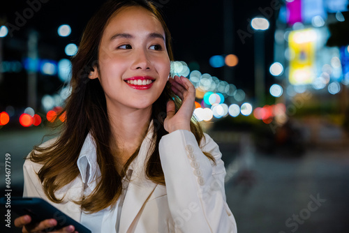 Smiling woman enjoying music through earphones attached to her mobile phone while strolling the night city streets. Wireless technology and digital devices make life convenient. Portrait of happy girl