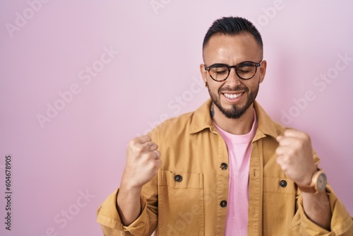 Young hispanic man standing over pink background very happy and excited doing winner gesture with arms raised, smiling and screaming for success. celebration concept. © Krakenimages.com