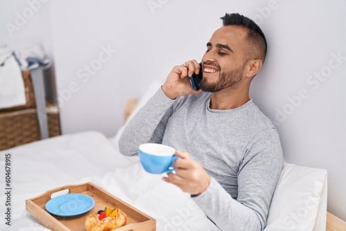 Young hispanic man talking on smartphone having breakfast at bedroom