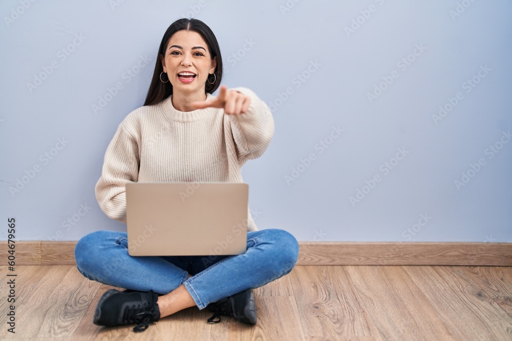 Young woman using laptop sitting on the floor at home laughing at you, pointing finger to the camera with hand over body, shame expression Young woman using laptop sitting on the floor at home laughing at you, pointing finger to the camera with hand over body, shame expression