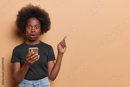 Horizontal shot of impressed black woman with bushy hair holds mobile phone reac Fototapet