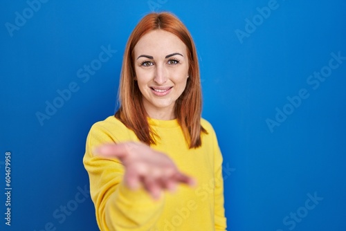 Young woman standing over blue background smiling cheerful offering palm hand giving assistance and acceptance.