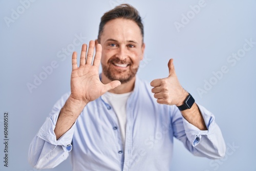 Middle age caucasian man standing over blue background showing and pointing up with fingers number six while smiling confident and happy. © Krakenimages.com