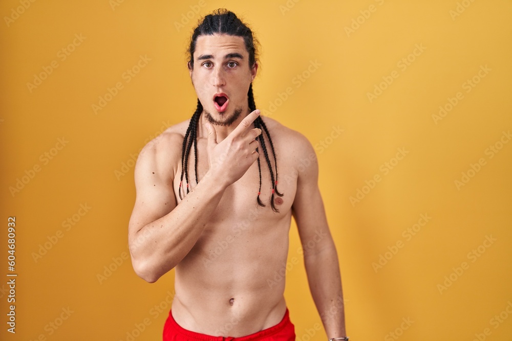 Hispanic man with long hair standing shirtless over yellow background looking fascinated with disbelief, surprise and amazed expression with hands on chin