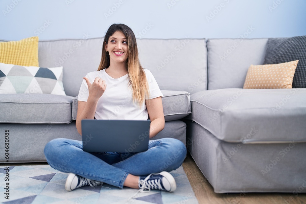 Young brunette woman sitting on the floor at home using laptop pointing thumb up to the side smiling happy with open mouth Young brunette woman sitting on the floor at home using laptop pointing thumb up to the side smiling happy with open mouth
