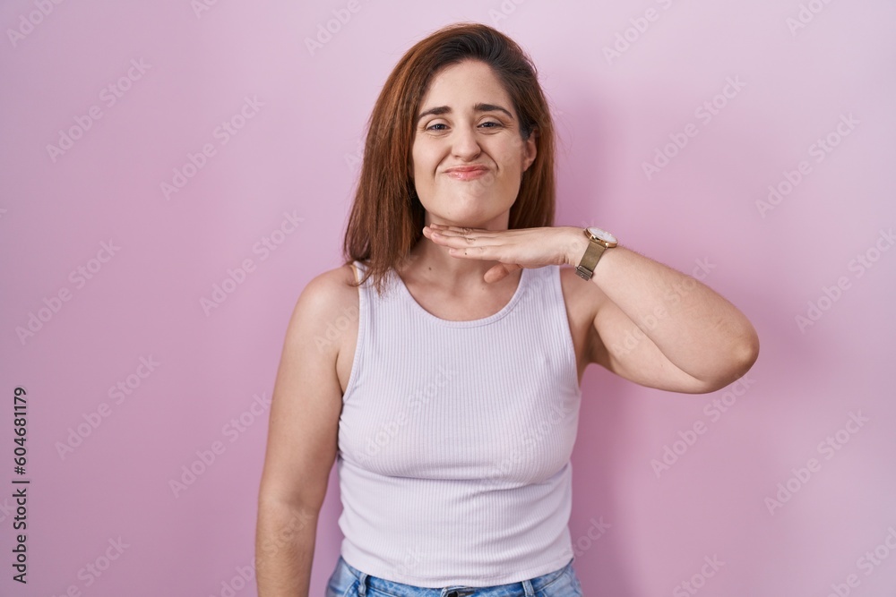 Brunette woman standing over pink background cutting throat with hand as knife, threaten aggression with furious violence Brunette woman standing over pink background cutting throat with hand as knife, threaten aggression with furious violence