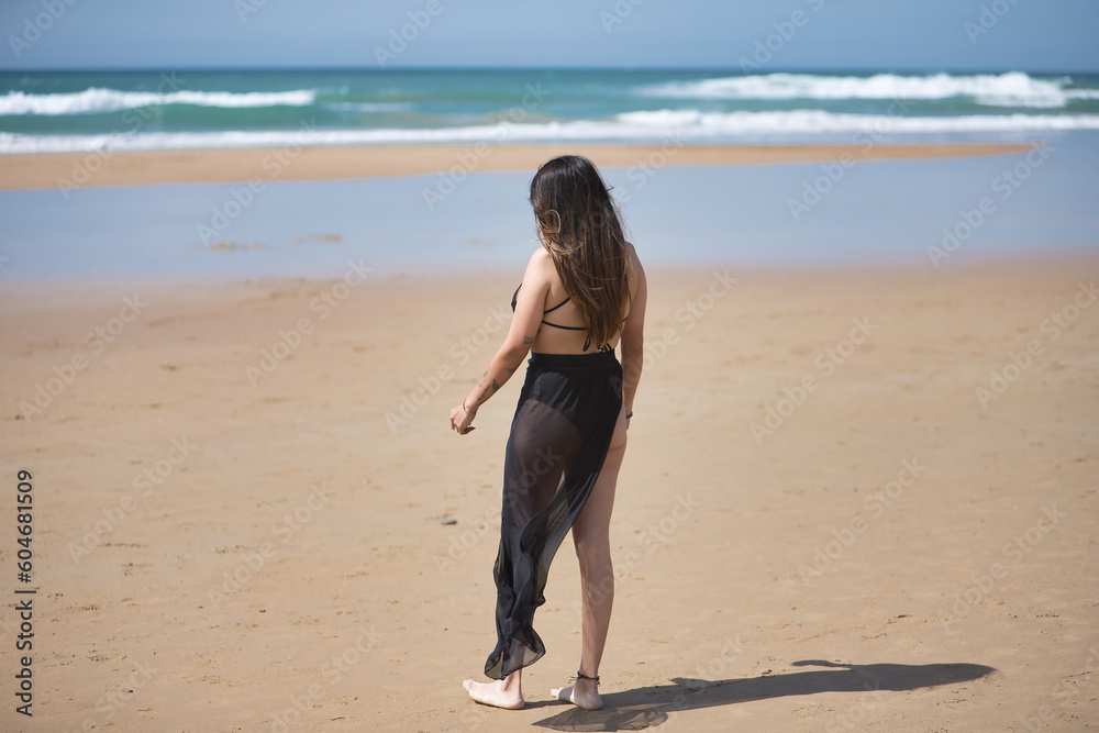 Attractive young woman in black swimsuit and sarong, solitary and quiet, walking on the beach, seen from the back. Concept tranquility, peace, wind, travel, vacation. Attractive young woman in black swimsuit and sarong, solitary and quiet, walking on the beach, seen from the back. Concept tranquility, peace, wind, travel, vacation.