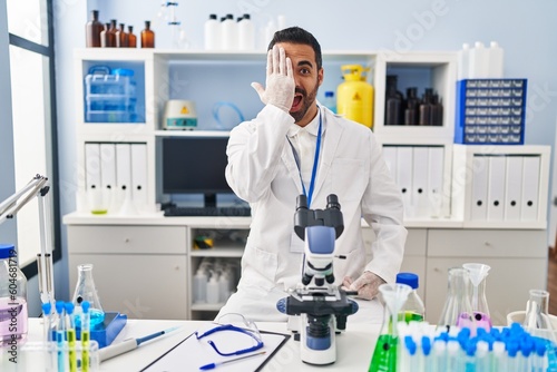 Young hispanic man with beard working at scientist laboratory covering one eye with hand, confident smile on face and surprise emotion.