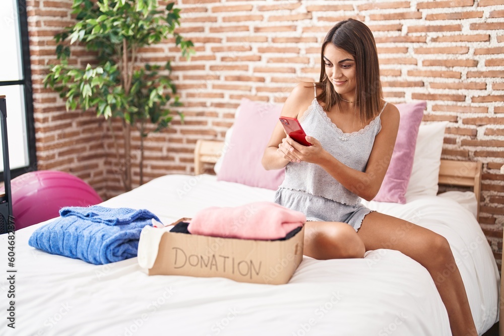 Young beautiful hispanic woman make photo to donation cardboard box with clothes at bedroom Young beautiful hispanic woman make photo to donation cardboard box with clothes at bedroom