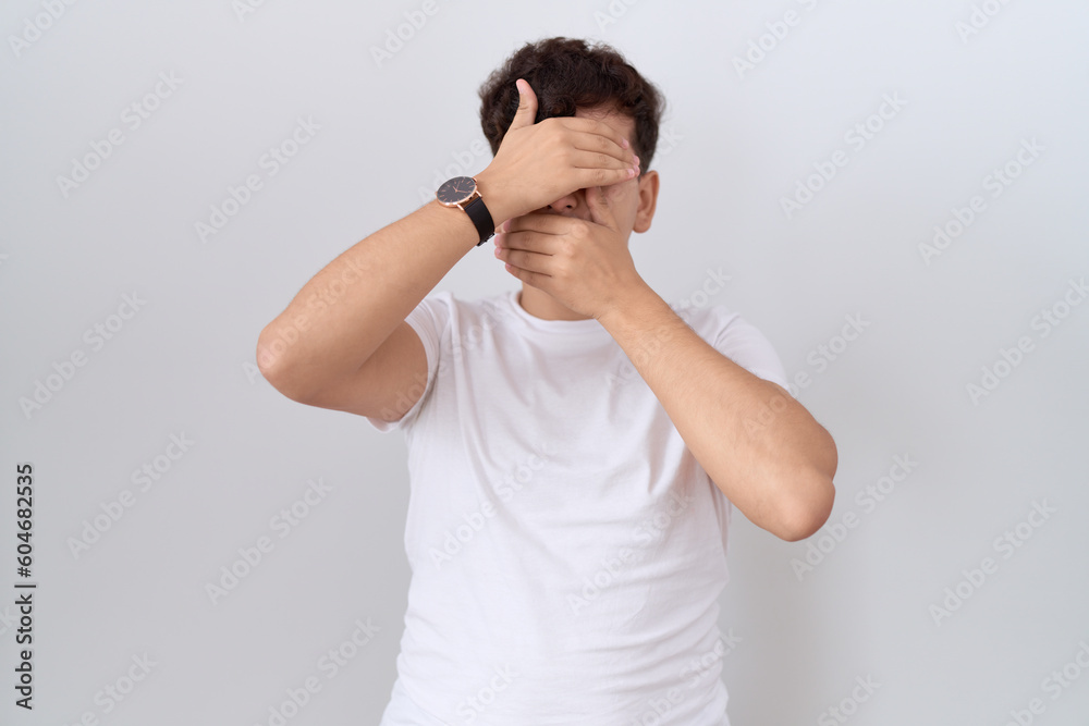 Young non binary man wearing casual white t shirt covering eyes and mouth with hands, surprised and shocked. hiding emotion Young non binary man wearing casual white t shirt covering eyes and mouth with hands, surprised and shocked. hiding emotion