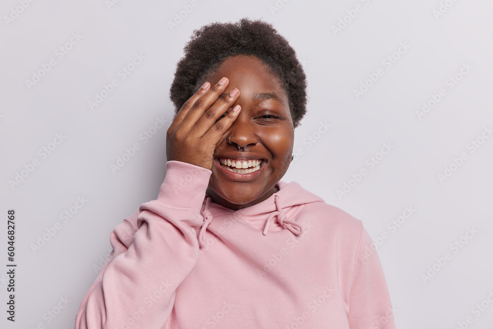 Overjoyed black woman makes face palm giggles positively smiles broadly laughs at something funny dressed in casual pink sweatshirt isolated over white background. People and emotions concept Overjoyed black woman makes face palm giggles positively smiles broadly laughs at something funny dressed in casual pink sweatshirt isolated over white background. People and emotions concept