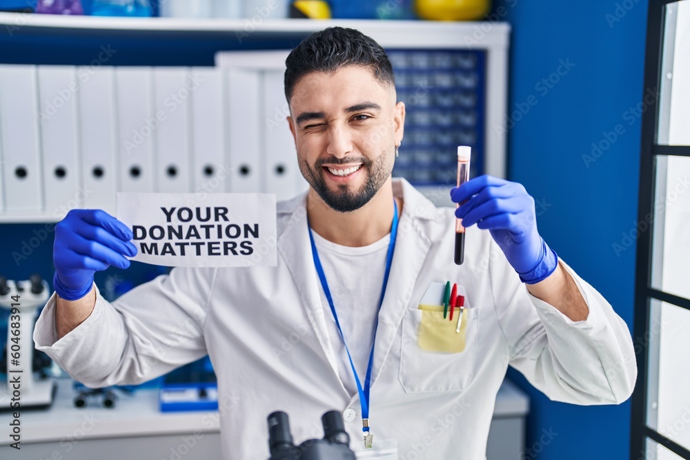 Young handsome man working at scientist laboratory holding blood sample winking looking at the camera with sexy expression, cheerful and happy face. Young handsome man working at scientist laboratory holding blood sample winking looking at the camera with sexy expression, cheerful and happy face.