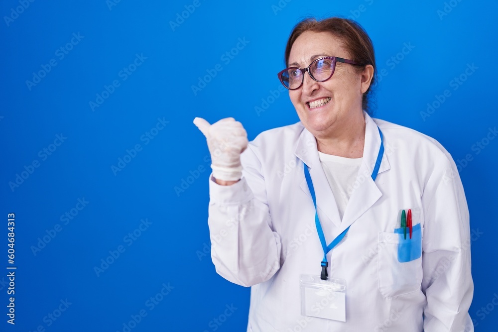 Senior woman with glasses wearing scientist uniform smiling with happy face looking and pointing to the side with thumb up.