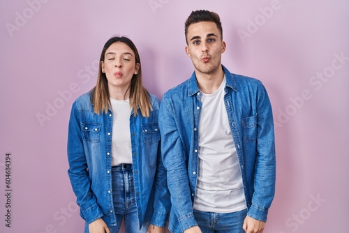 Young hispanic couple standing over pink background making fish face with lips, crazy and comical gesture. funny expression.