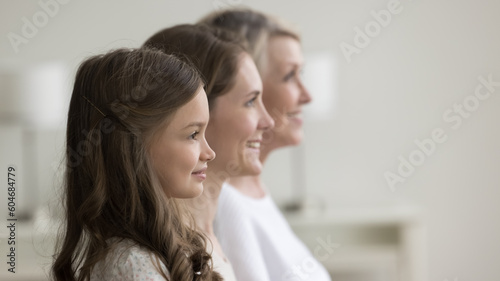 Side profile faces view of happy three generation of relatives women. Beautiful preschooler girl looking forward standing posing indoors in row with young pretty mother and senior cheerful grandmother