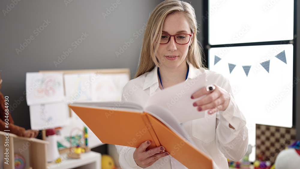 Young blonde woman preschool teacher smiling confident reading book at kindergarten Young blonde woman preschool teacher smiling confident reading book at kindergarten