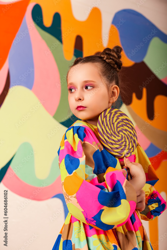 Scared little girl posing with round large lollipop at multi colours wall backdrop, pensive looking away. Lovely kid in colorful dress holding candy on stick. Summer sweet concept. Copy ad text space