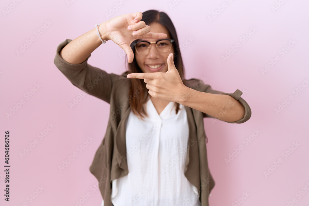 Middle age chinese woman wearing glasses over pink background smiling making frame with hands and fingers with happy face. creativity and photography concept. Middle age chinese woman wearing glasses over pink background smiling making frame with hands and fingers with happy face. creativity and photography concept.