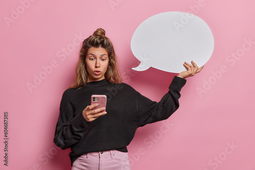 Stunned speechless young woman stares in smartphone with omg expression wears casual black jumper and trousers holds blank speech bubble isolated over pink background. Human reactions concept