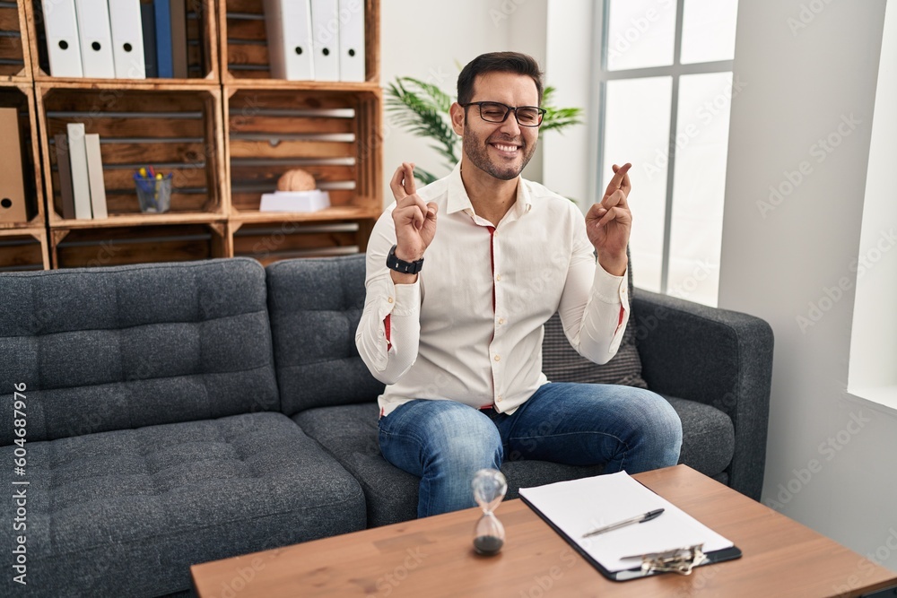 Young hispanic man with beard working at consultation office gesturing finger crossed smiling with hope and eyes closed. luck and superstitious concept. Young hispanic man with beard working at consultation office gesturing finger crossed smiling with hope and eyes closed. luck and superstitious concept.