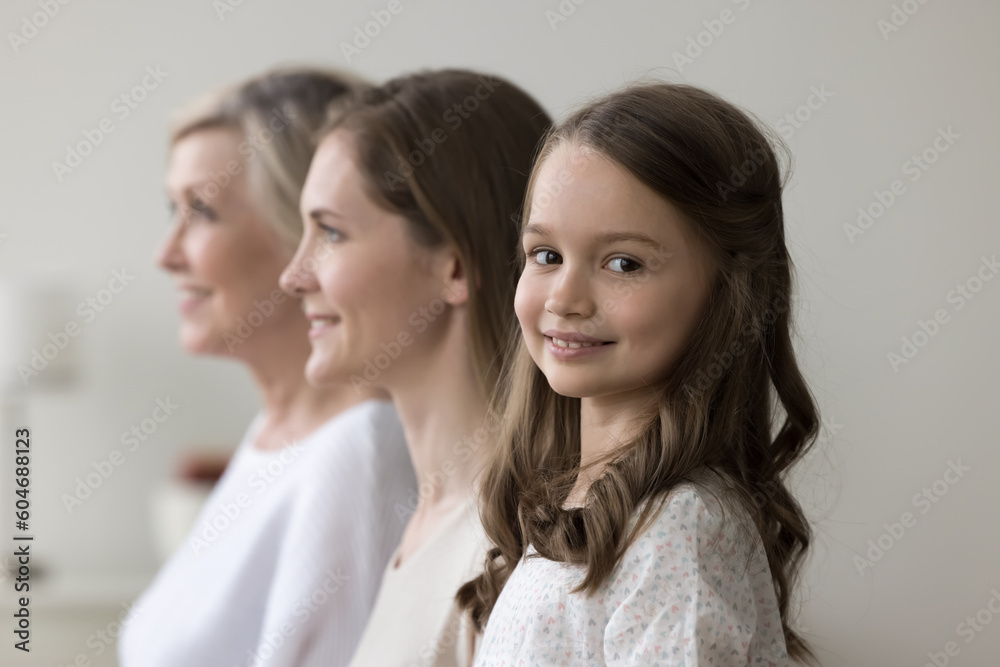 Happy preschooler girl staring at camera posing standing in row with young pretty mother and older grandmother look forward. Multigenerational family shooting indoor. Life from childhood to retirement Happy preschooler girl staring at camera posing standing in row with young pretty mother and older grandmother look forward. Multigenerational family shooting indoor. Life from childhood to retirement