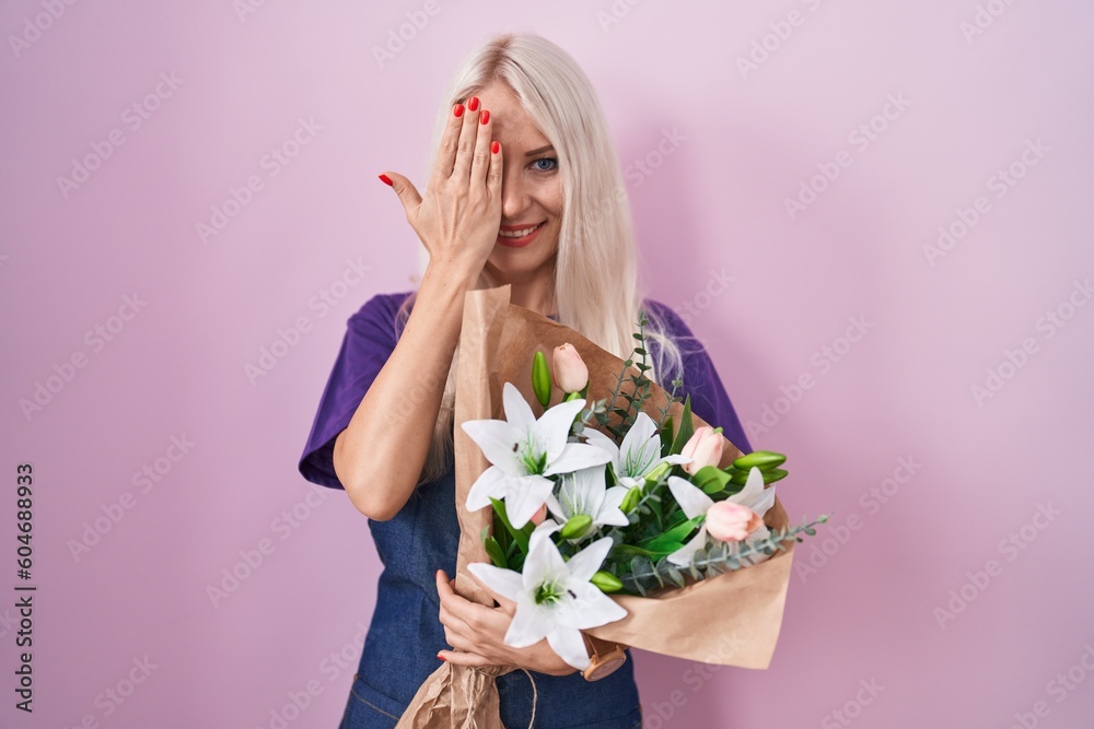 Caucasian woman holding bouquet of white flowers covering one eye with hand, confident smile on face and surprise emotion.