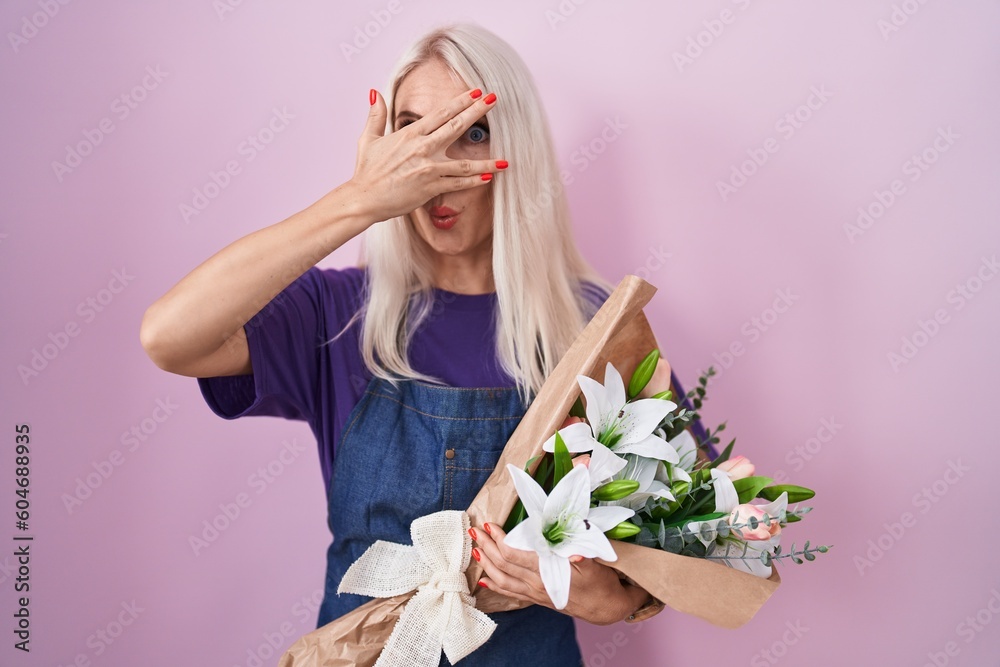 Caucasian woman holding bouquet of white flowers peeking in shock covering face and eyes with hand, looking through fingers with embarrassed expression.