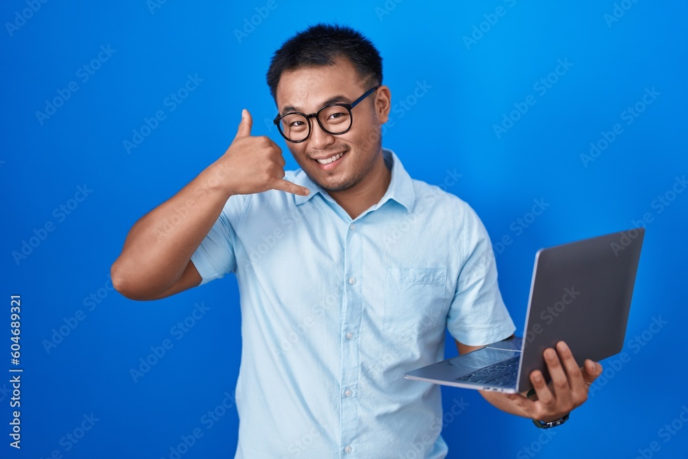 Chinese young man using computer laptop smiling doing phone gesture with hand and fingers like talking on the telephone. communicating concepts. Chinese young man using computer laptop smiling doing phone gesture with hand and fingers like talking on the telephone. communicating concepts.