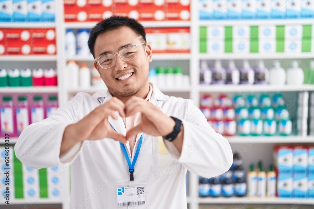 Chinese young man working at pharmacy drugstore smiling in love doing heart symbol shape with hands. romantic concept. Chinese young man working at pharmacy drugstore smiling in love doing heart symbol shape with hands. romantic concept.