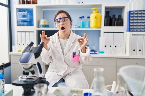 Hispanic girl with down syndrome working at scientist laboratory looking surprised and shocked doing ok approval symbol with fingers. crazy expression