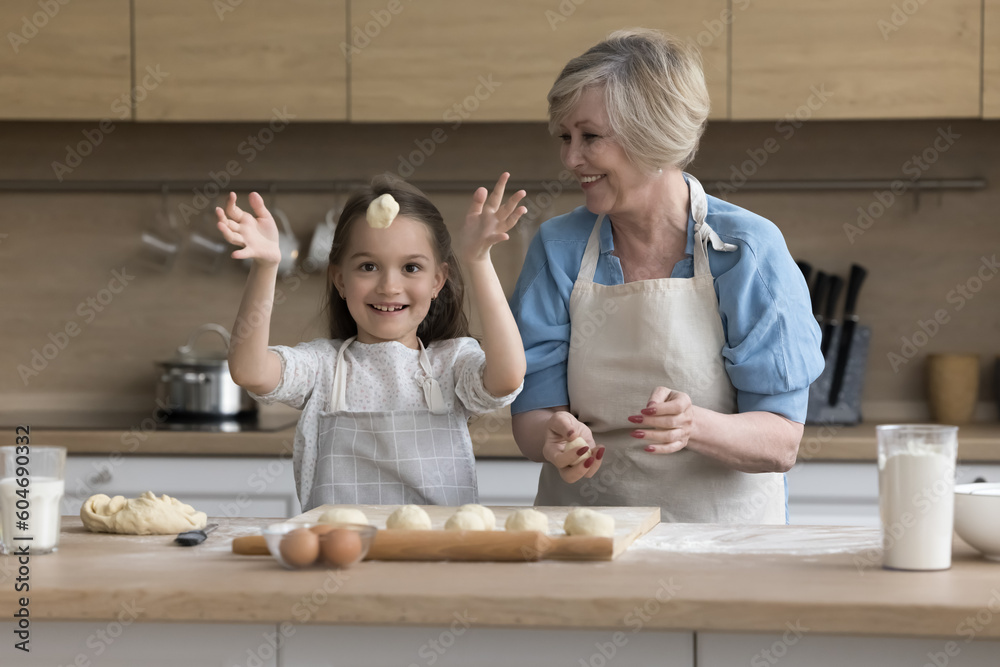 Attractive retired granny teach cute funny granddaughter to cook, prepare buns or cookies, make handmade pastries, happy child throwing dough looks playful white cooking with loving grandma in kitchen